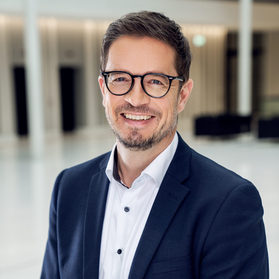 Stellvertretender Pressesprecher Matthias Eichler lächelt in die Kamera, er trägt eine Brille mit rundem Rand, einen Bart und ein dunkelblaues Jackett