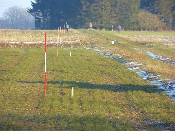 Menschen messen an an einem Feldrand die Wegesgrenzen. Es ist Winter, es ist Schnee zu sehen.