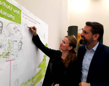 Minister Olaf Lies steht gemeinsam mit einer Zeichnerin vor einem großen Plakat, die ein Bild von dem Minister skizziert.