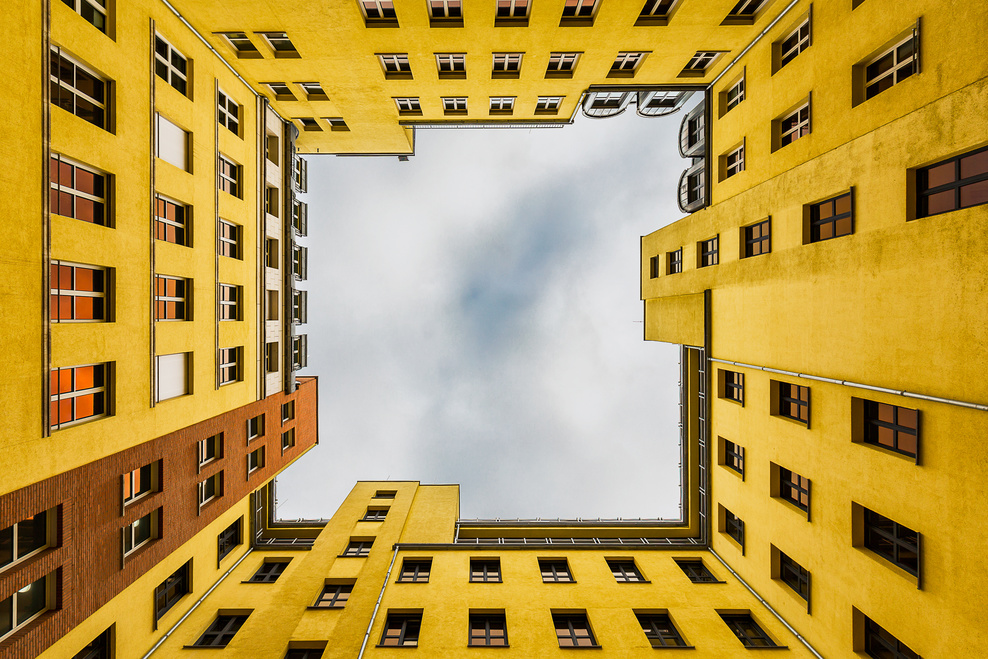Zu sehen ist ein gelber Wohnblock mit mehreren Fenstern. Der Betrachter steht im Innenhof und schaut nach oben entlang der Fassaden in den wolkenbehangenen Himmel.