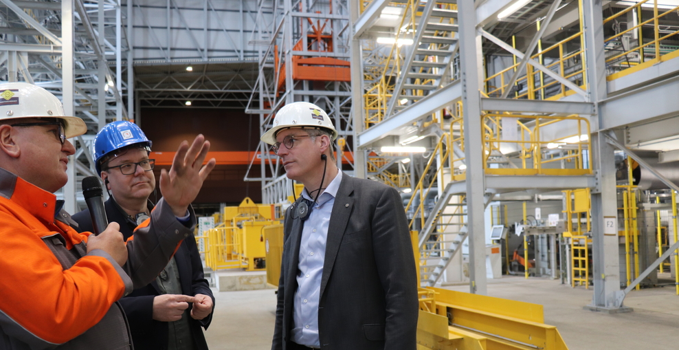 Minister Meyer bei seinem Besuch bei der Salzgitter AG, er steht in einer Fabrikhalle, trägt einen blauen Arbeitshelm und weitere Schutzkleidung, ein Mitarbeiter erklärt ihm gestikulierend, was er gerade sieht