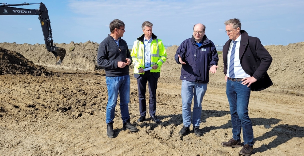 Christian Meyer und Mitarbeiter des NLWKNs stehen auf der Baustelle einer Deicherhöhung, im Hintergrund sieht man Sand und den Arm eines Baggers