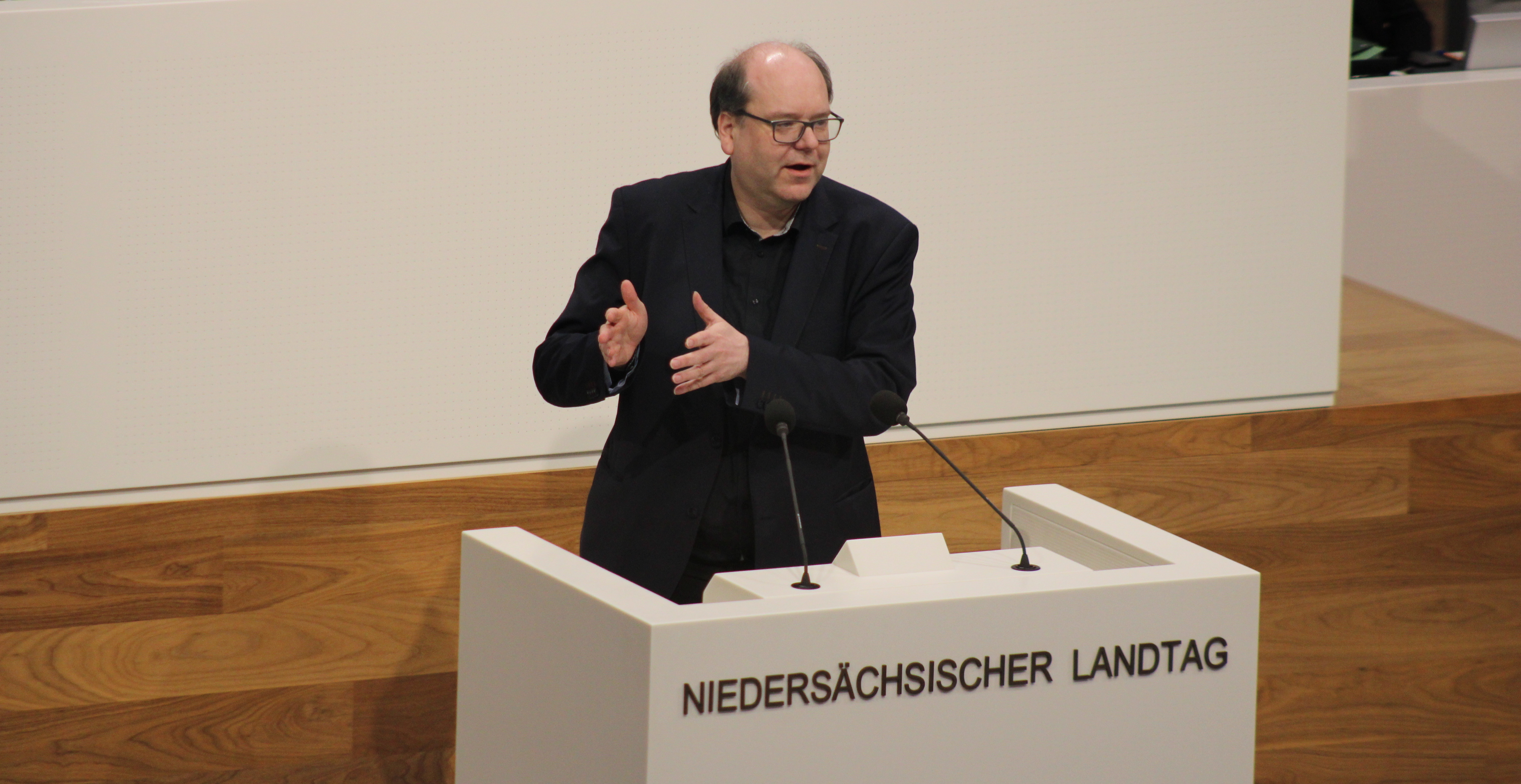 Minister Christian Meyer am Rednerpult im niedersächsischen Landtag. Er trägt einen dunkelblauen Pullover, ein dunkles Jackett und hat die Arme angewinkelt, um zu gestikulieren