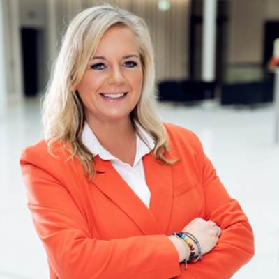 Redaktionsassitentin Marita Haouari steht mit verschränkten Armen leicht seitlich zur Kamera und lächelt, sie hat blondes Haar, das über die Schultern reicht und trägt einen orangenen Blazer und mehrere Armbänder